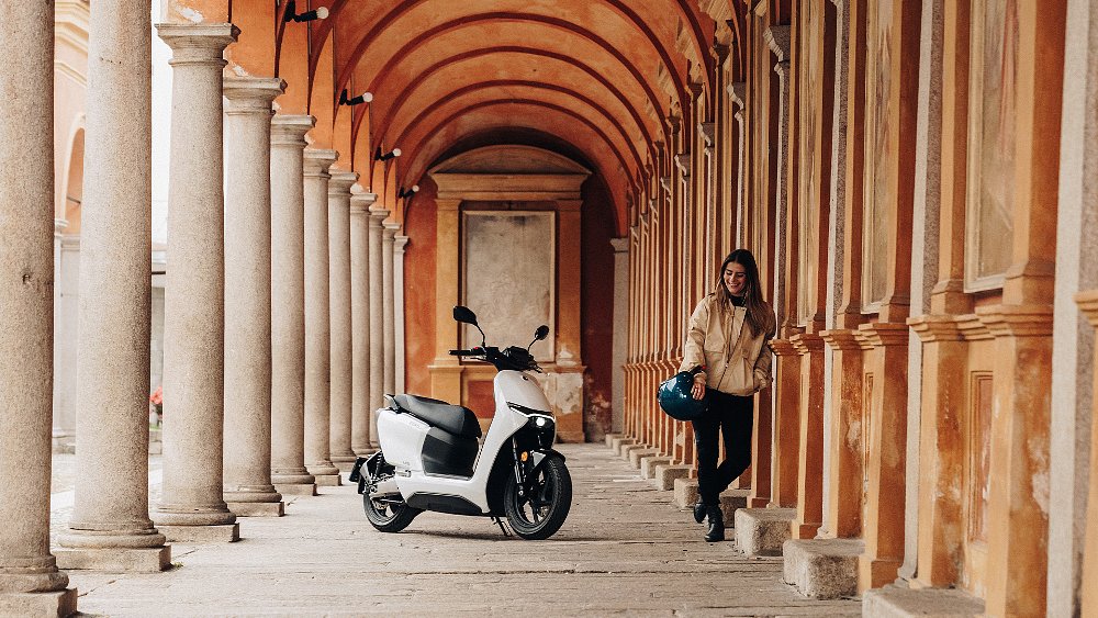 A female rider stands beside the Zero LS1 electric scooter in a column-lined corridor.