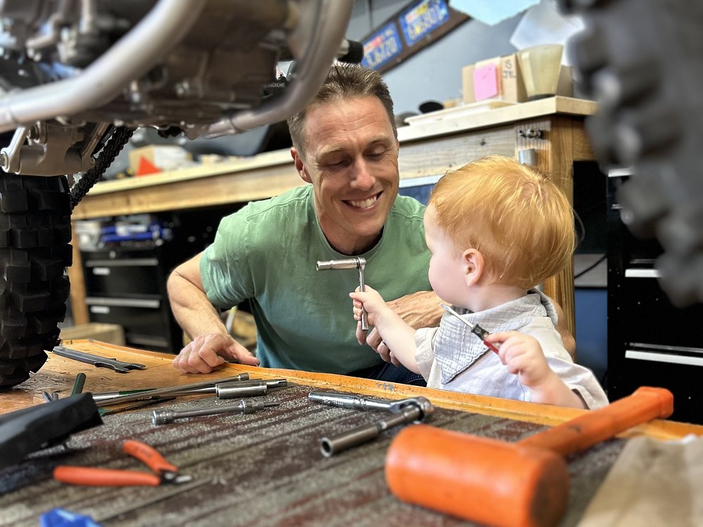 Ari in the garage with a motorcycle partly disassembled and young Milo holding a tool