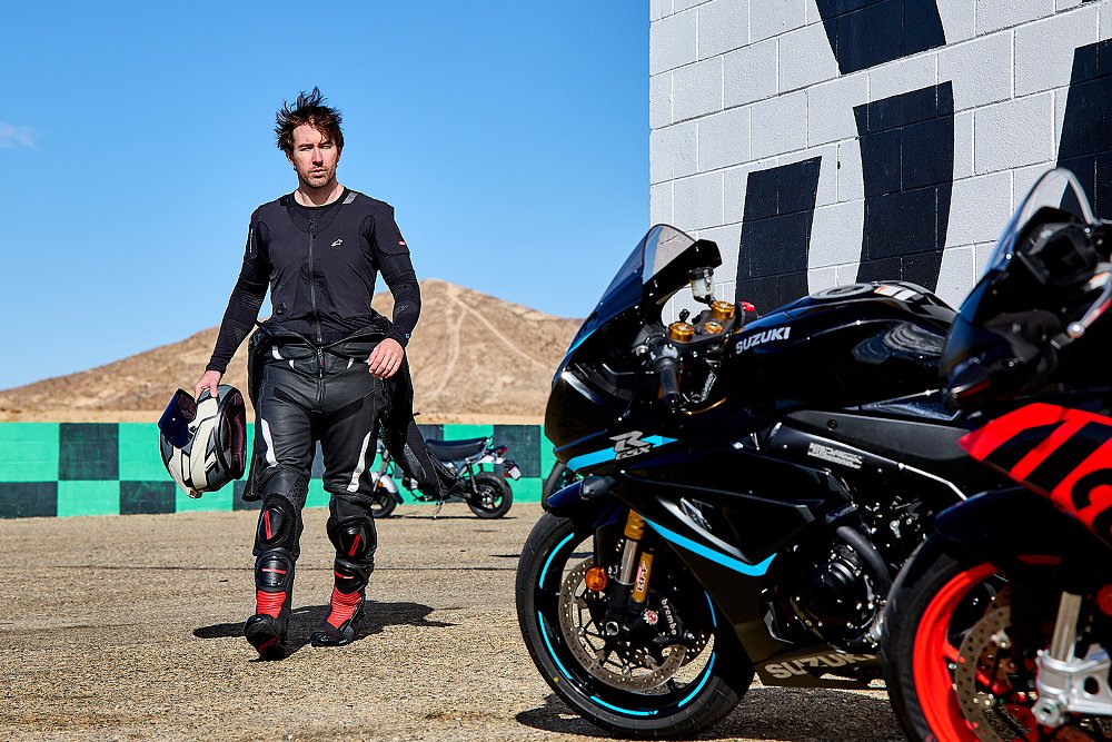 A track rider, outfitted in a race suit and airbag, walks toward two sport bikes.