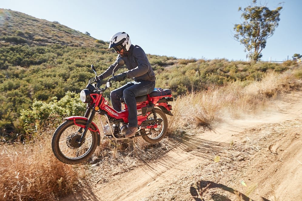 Zack riding the red Honda Trail 125 on a dirt road, flying over a small jump