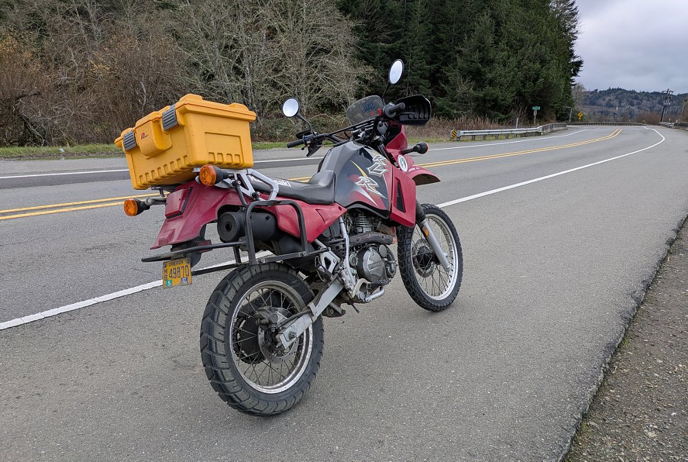 the red KLR650 with a large yellow box on the rear rack, parked along a forest road