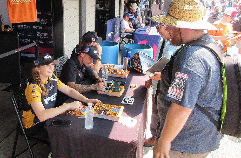 three riders at a table signing posters for fans