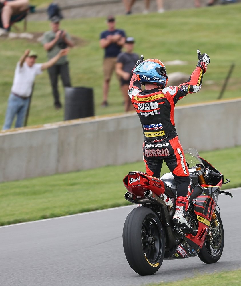 Josh Herrin celebrates a race win at Road America, standing on his motorcycle with thumbs up as fans cheer