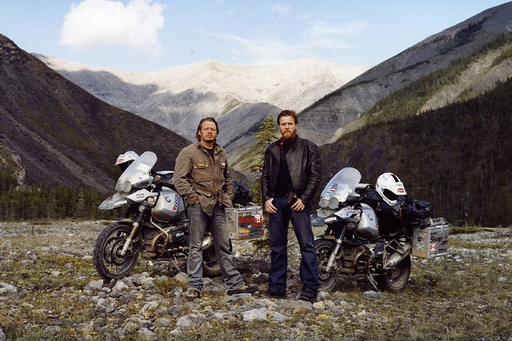 Charley Boorman and Ewan McGregor pose by their motorcycles with mountains in the background.