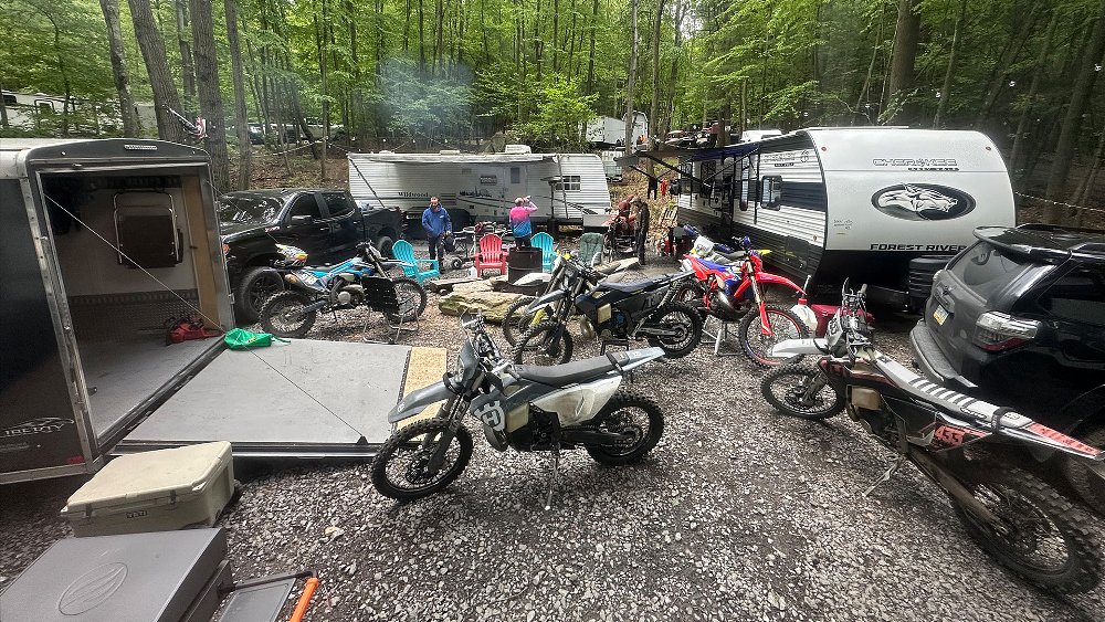 A collection of trailers, trucks, dirt bikes, and riders gather around a campsite.