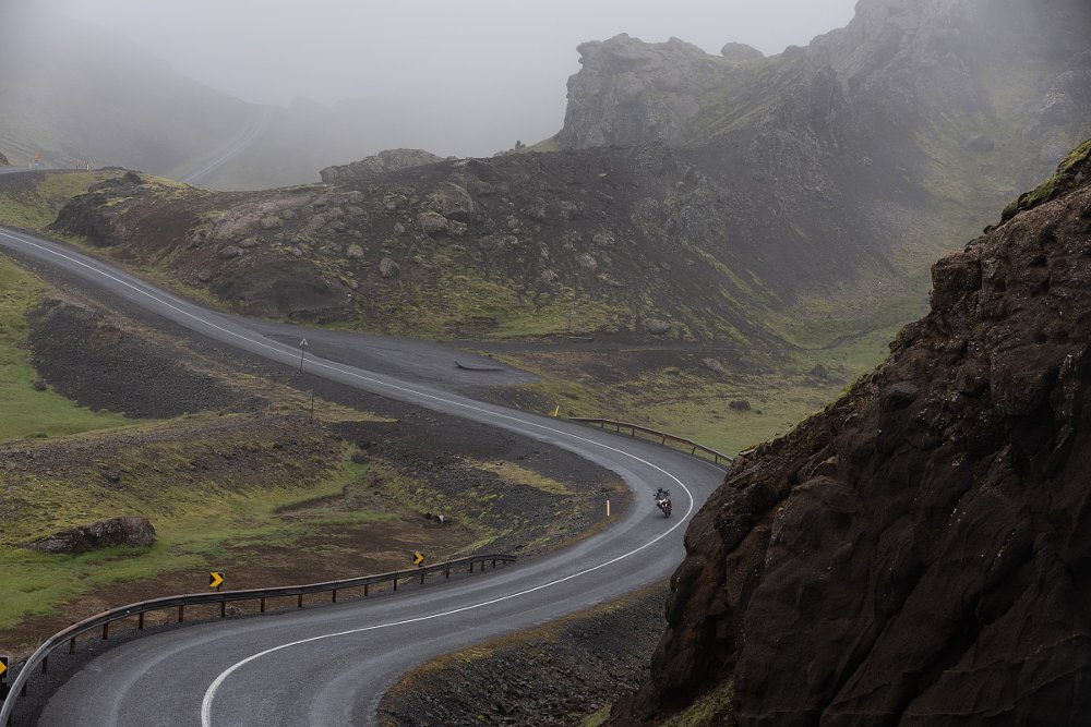 long view of the motorcycle riding on a curving, wet road under cloudy skies and between mountains
