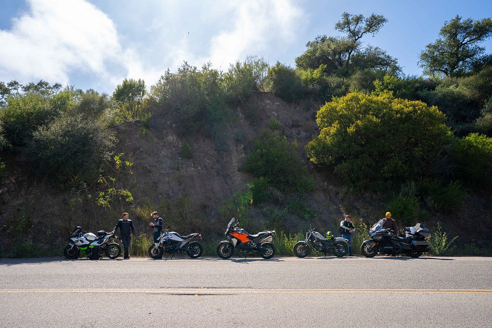 Five motorcycles parked next to a road with riders talking in the background.