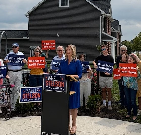 Janelle Stelson making a campaign speech with supporters behind her, some holding signs saying 'Repeal the Tariff Taxes'