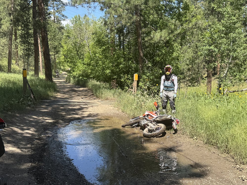 A 2026 KTM 690 Enduro R tipped over next to a puddle on a dirt road, with a rider standing next to it.