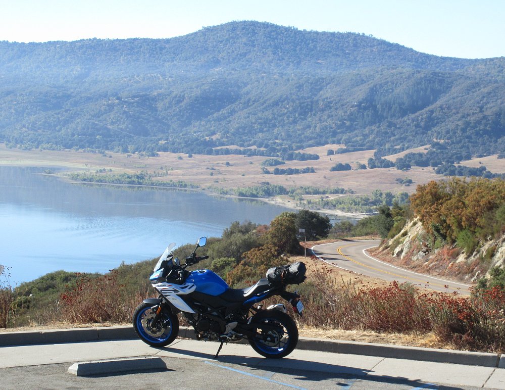the SV-7GX at a mountain overlook, curving road in the background, above a lake, a bag strapped on the motorcycle