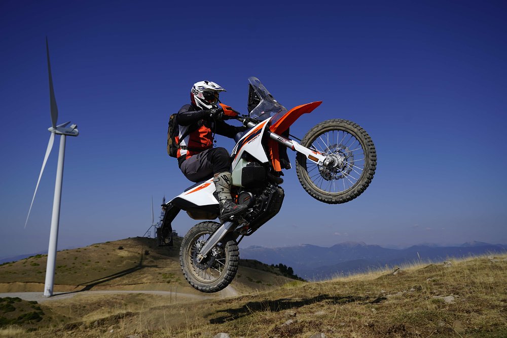 2027 KTM 690 Rally jumping off a small hillside with a large windmill in the background.