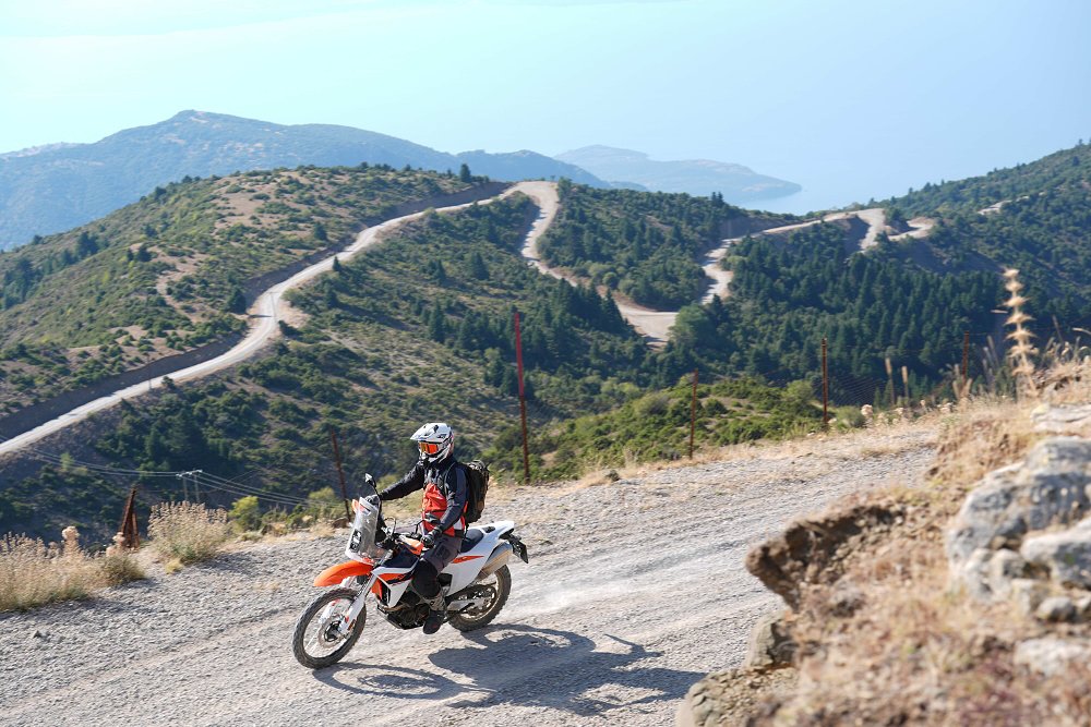 2027 KTM 690 Rally riding on a dirt road with trails and hills in the background.