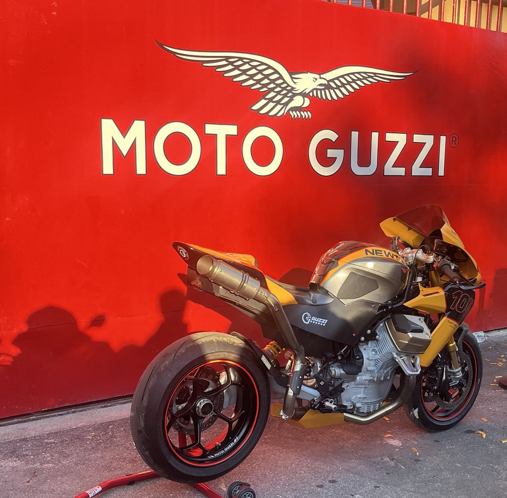 gold and silver V-twin sport bike with single-sided swingarm in front of the red gate at the factory entrance
