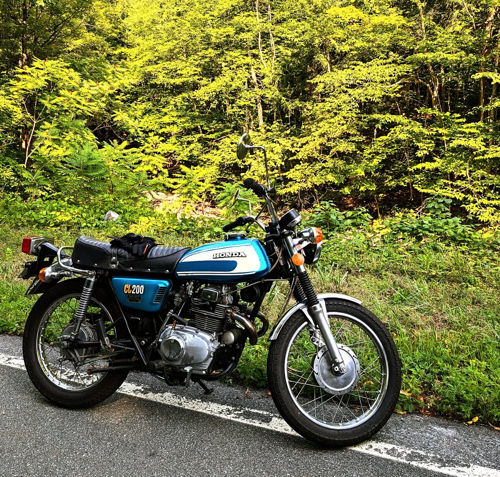 the blue CL200 Scrambler parked in front of sunlit trees alongside a road