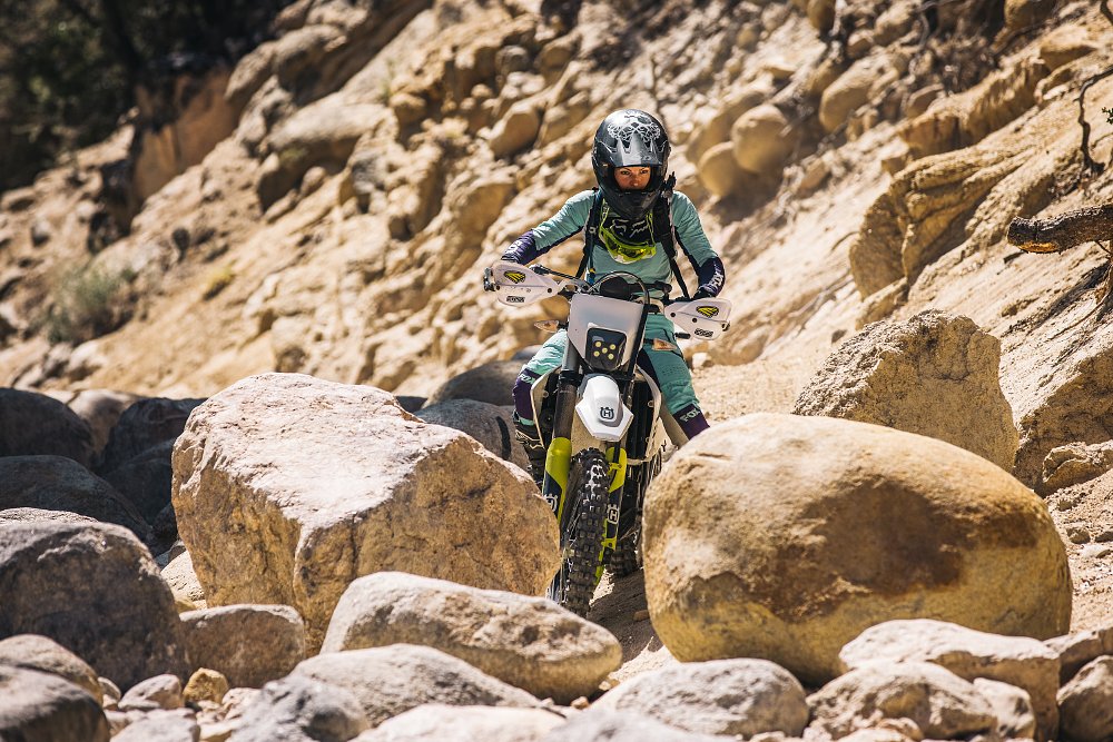A female rider riding inbetween very large boulders on board a Husqvarna dual sport motorcycle.
