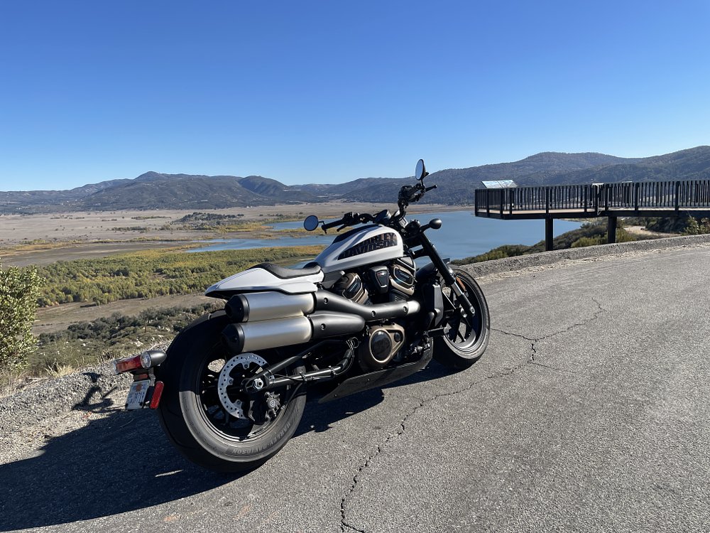 A white Harley-Davidson sportster s sits on a lakeside vista, rear three quarter view with big exhaust pipes