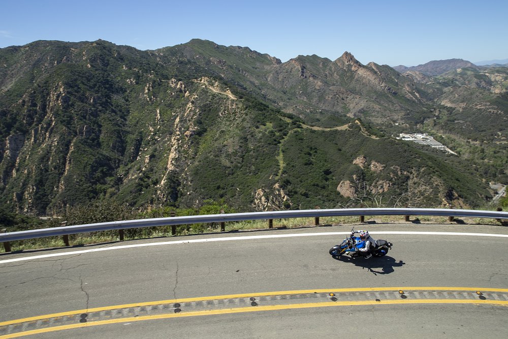 A wide view of a mountain cliffside road with the GSX-S1000 leaned over in the corner riding through