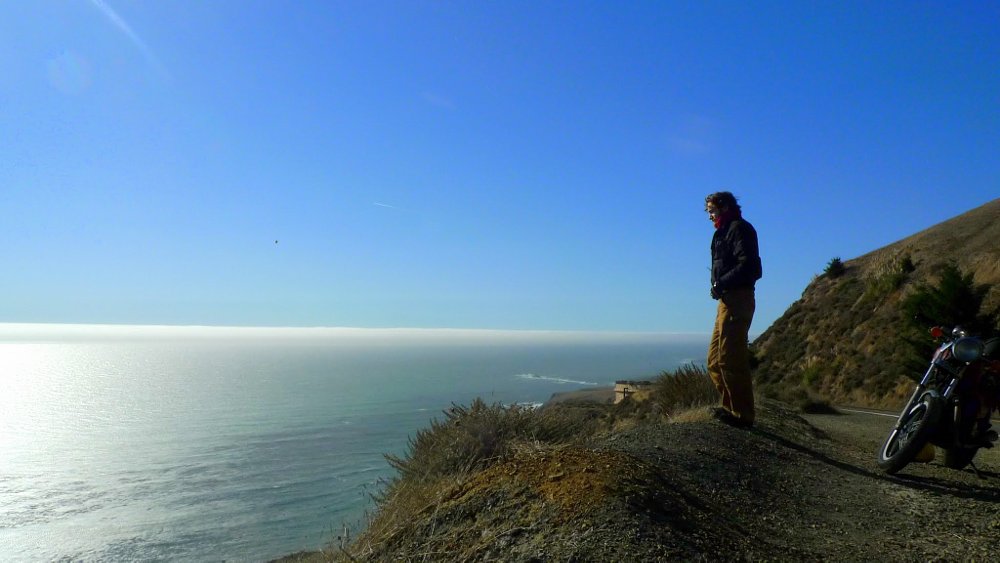 rider on a mountainside overlooking the ocean with the motorcycle parked nearby