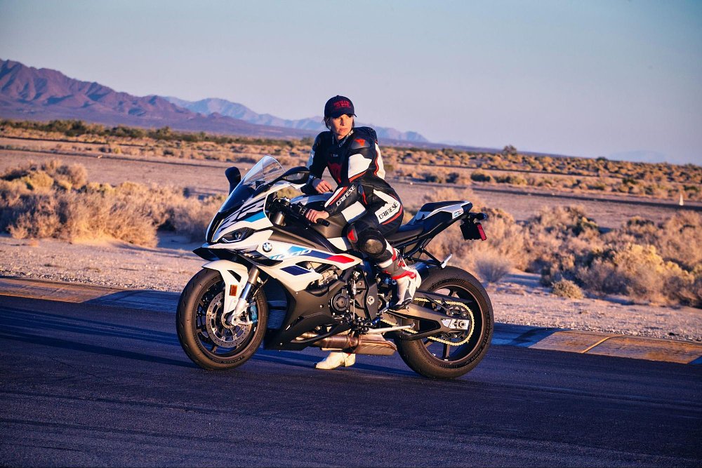 A female rider poses on top of the BMW S 1000 RR in full race leathers on the race track