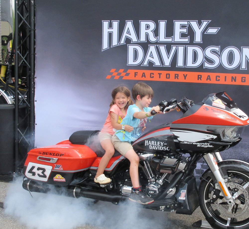 small boy and girl smiling while on a Harley-Davidson race bike with smoke pouring from the rear wheel