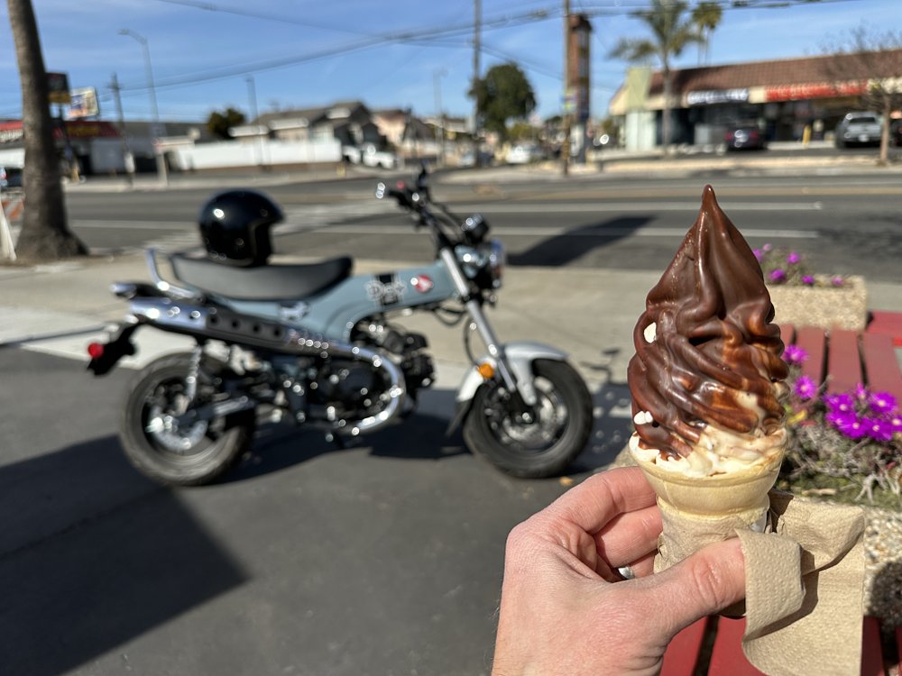 A chocolate-dipped soft serve ice cream with a 2025 Honda Dax 125 in the background.