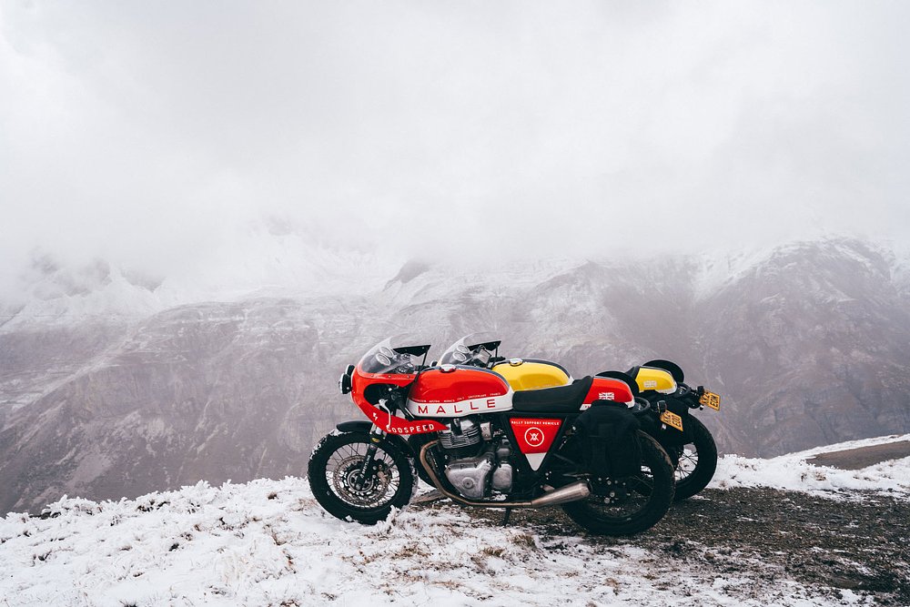one red and one yellow custom Royal Enfield fitted with luggage on a snowy mountainside