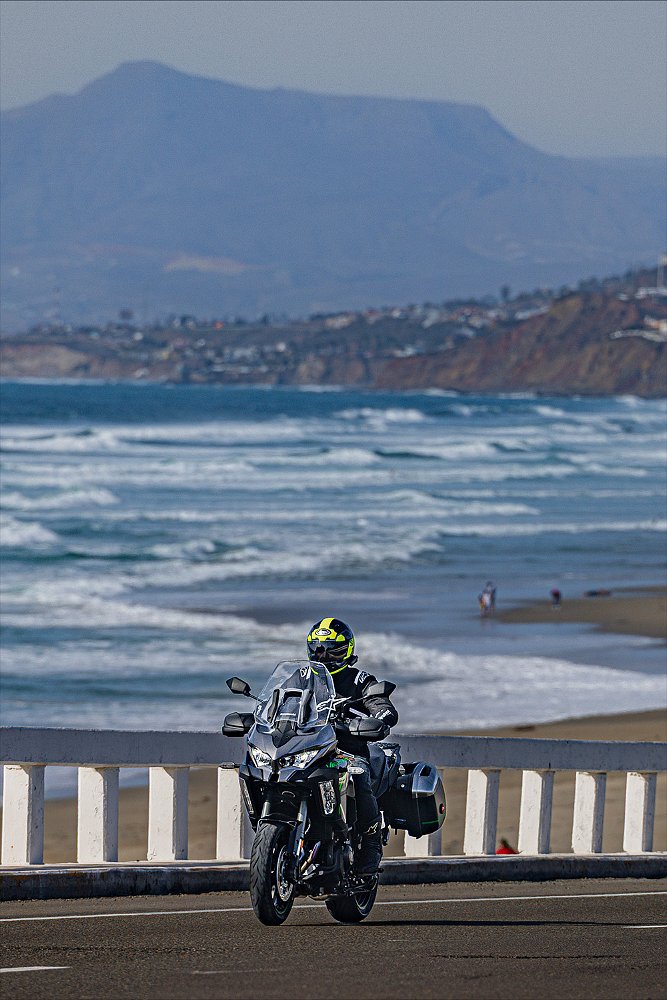 Dustin rides the Versys 1100 SE LT with waves crashing on the beach in the background.