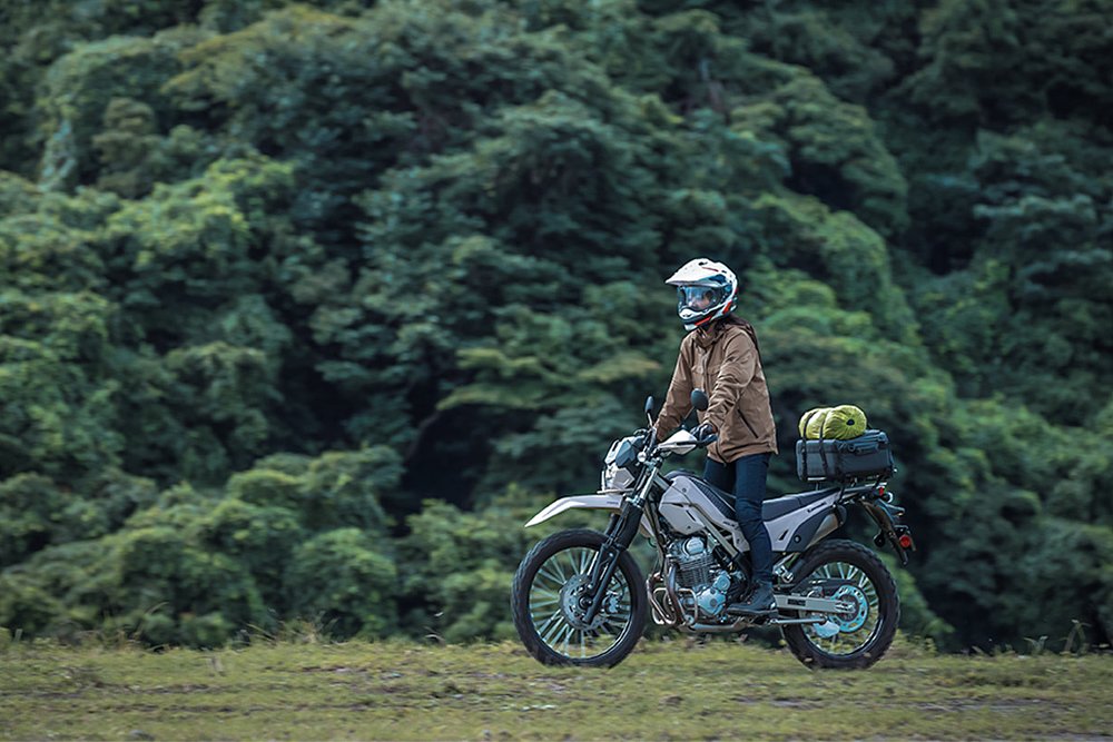 A KLX230 Sherpa S rider explores a green meadow while standing on the bike's footpegs.