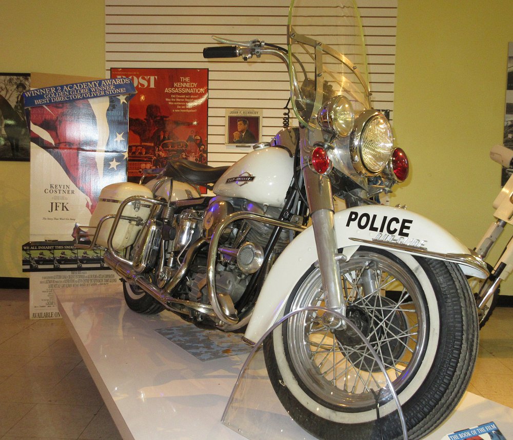 A white police motorcycle from the 1960s in front of a 'JFK' movie poster