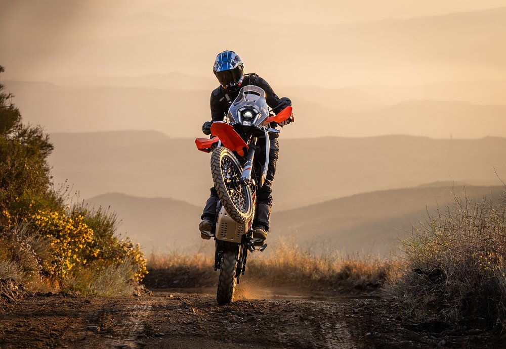 2025 KTM 390 Adventure R wheelie toward camera with a sunset behind.