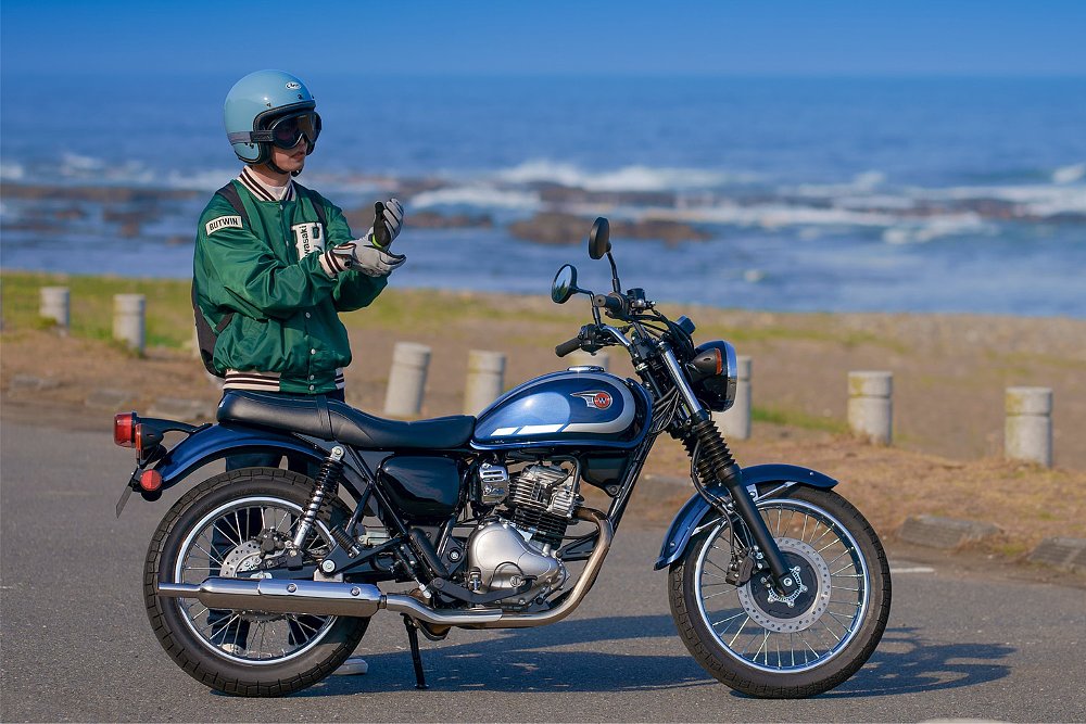A rider stands next to the Kawasaki W230 retro roadster with a beach in the background.