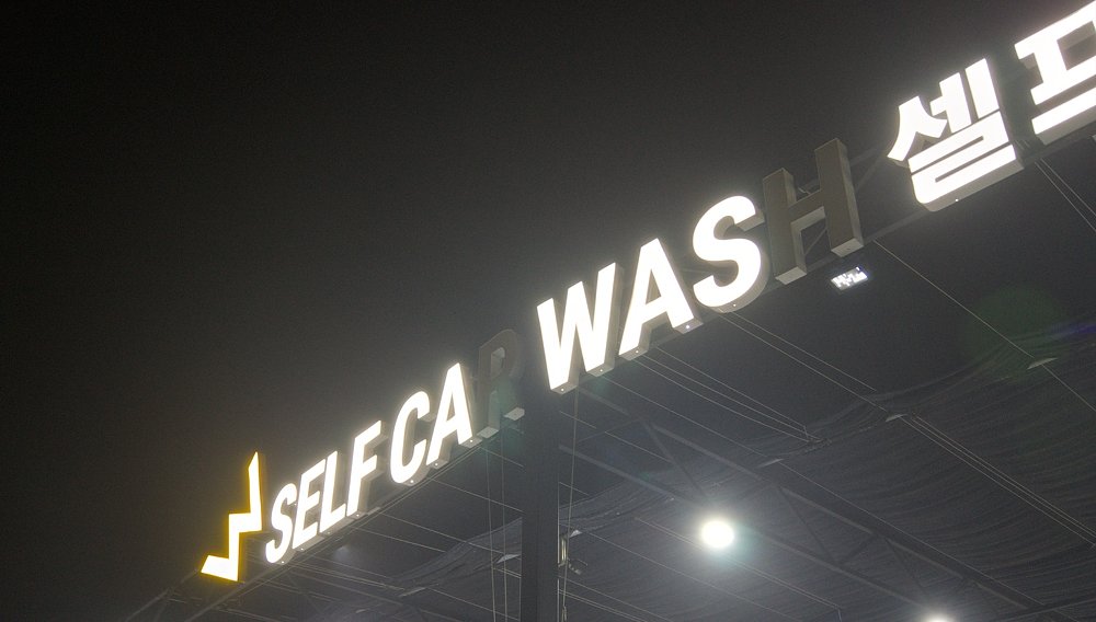 illuminated 'self car wash' sign in English and Korean with some letters burned out, surrounded by fog