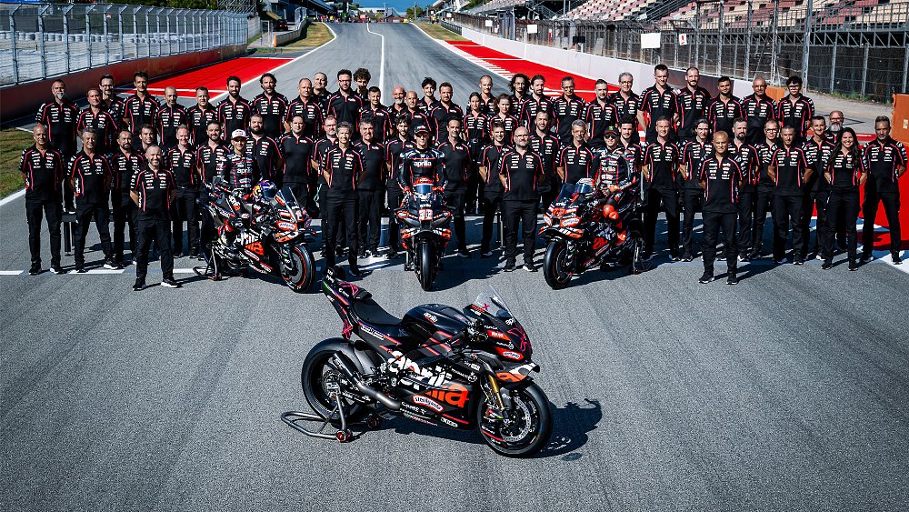 The Aprilia RSV4 X-GP stands on the racetrack with the Aprilia MotoGP team in the background.