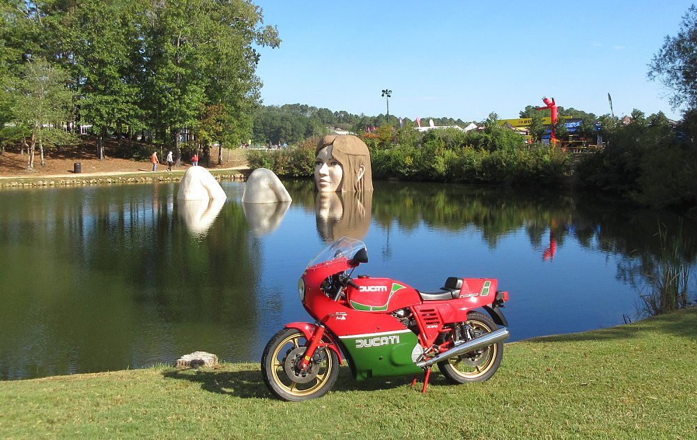 vintage red and green Ducati parked in front of a lake at the track with a huge statue of a woman's head and knees, as if she's taking a bath in the lake