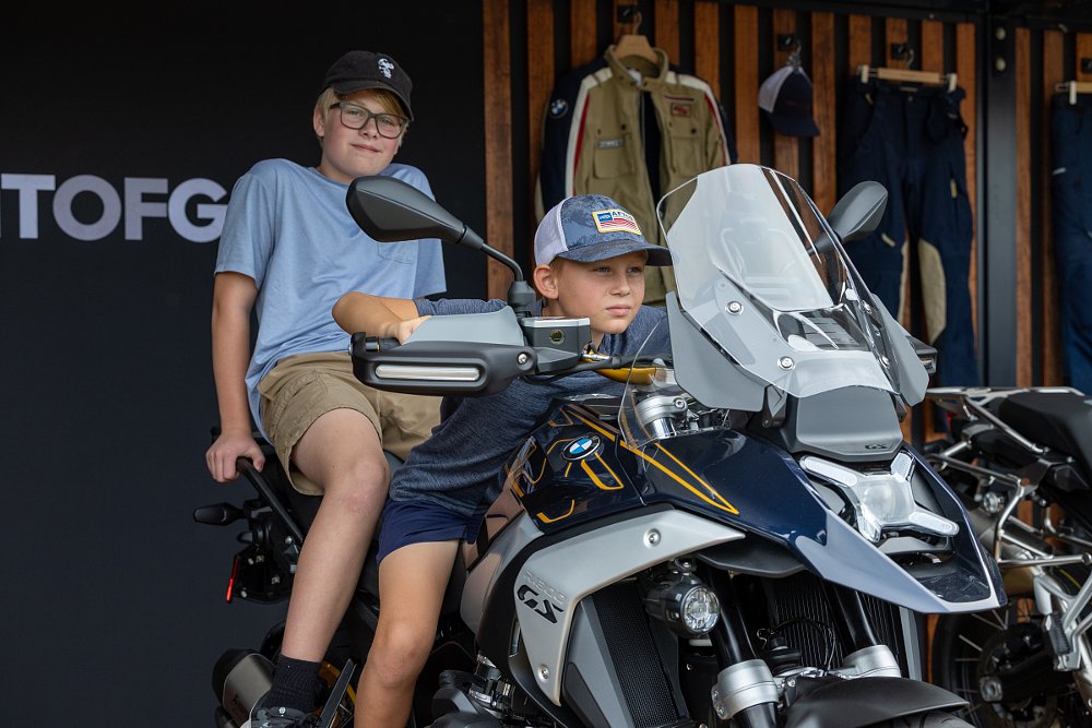 two young kids sitting on a new BMW motorcycle, the first one tucking in a pretending to ride fast